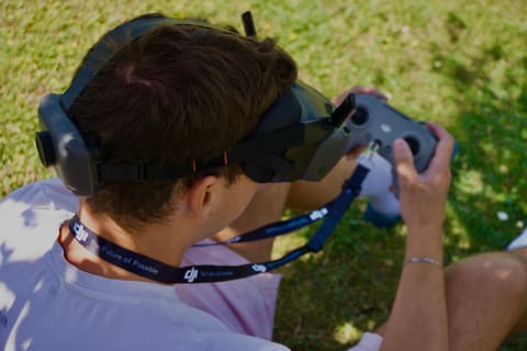 Pilote vu de dos, casque FPV sur les yeux et radiocommande en main, assis dans l'herbe pendant un vol.