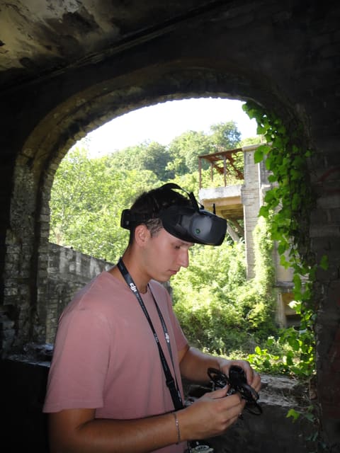 Préparation du drone sous une arche de pierre d'un bâtiment abandonné, casque FPV relevé sur le front.