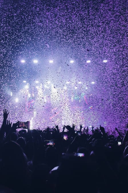Foule de concert bras levés sous une pluie de confettis, projecteurs violets braqués sur la scène.
