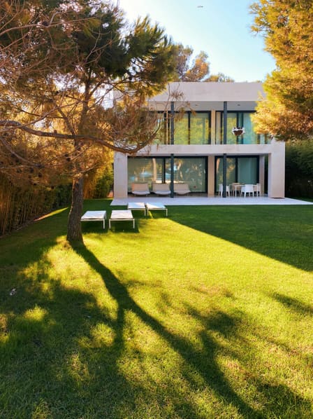 Villa contemporaine aux grandes baies vitrées, vue depuis la pelouse à l'ombre d'un pin.