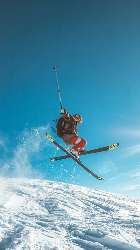 Skieur en plein saut au-dessus d'une pente enneigée, bâtons et skis croisés sur un ciel bleu.